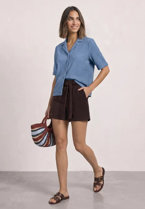 Frau trägt hellblaues kurzärmliges Hemd, braune Shorts, Sandalen und hält bunte Umhängetasche, lächelnd.