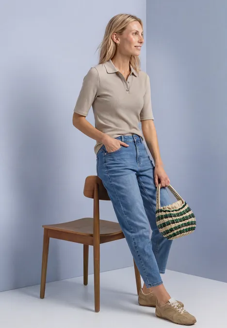 Poloshirt im Strick-Look mit Rippstruktur clay sand mel. Eine Frau in beigefarbenem Poloshirt, blauen Jeans und Loafers lehnt an einem Stuhl und hält eine Korbtasche.