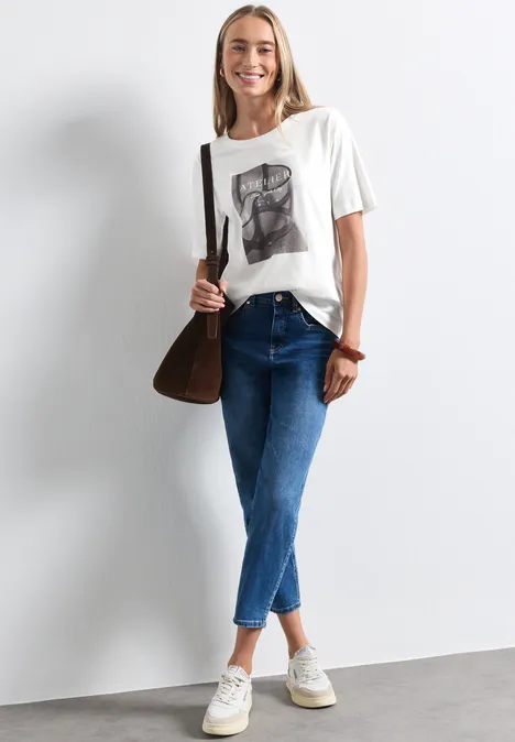 Shirt met ronde hals en fotoprint off white Eine junge Frau in Jeans, T-Shirt und Sneakers mit brauner Tasche lächelt vor weißem Hintergrund.