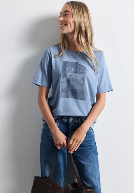 Shirt met ronde hals en fotoprint northern blue Eine lächelnde Frau in blauem T-Shirt und Jeans lehnt an einer weißen Wand und hält eine Tasche.