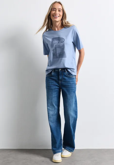 Shirt met ronde hals en fotoprint northern blue Eine lächelnde Frau in blauem T-Shirt, weiten Jeans und weißen Sneakern vor neutralem Hintergrund.