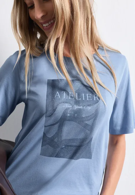 Shirt met ronde hals en fotoprint northern blue Eine lächelnde Frau trägt ein hellblaues T-Shirt mit grafischem Print 'Atelier'.
