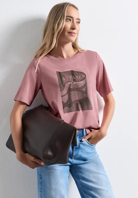 Shirt met ronde hals en fotoprint vintage blush Frau in rosafarbenem T-Shirt mit Print, blauen Jeans und großer brauner Tasche vor weißem Hintergrund.