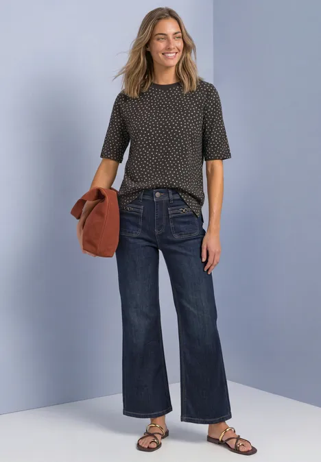 T-Shirt mit Rundhals und Print black coffee Frau mit gemustertem Shirt, weitem Jeans und Sandalen, hält eine Jacke vor hellgrau-blauem Hintergrund.