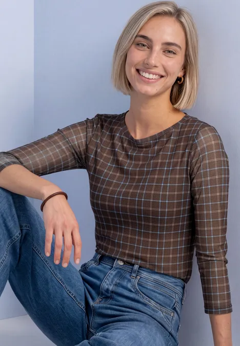 Shirt met 3/4-mouwen van mesh met print silk brown Lachende vrouw met kort blond haar, in een ruitjes top en spijkerbroek, zittend voor een lichte achtergrond.