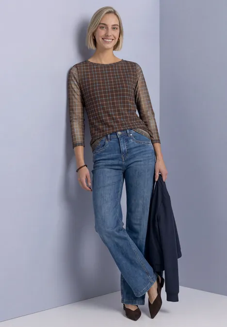 Shirt met 3/4-mouwen van mesh met print silk brown Vrolijke vrouw met kort blond haar in geruit shirt en blauwe jeans leunt tegen muur en houdt een jas vast.