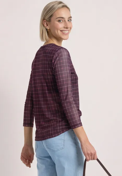 Shirt met 3/4-mouwen van mesh met print rustic rouge Eine lächelnde Frau in rotem, kariertem Langarmshirt und hellblauen Jeans, die eine kleine Handtasche hält.