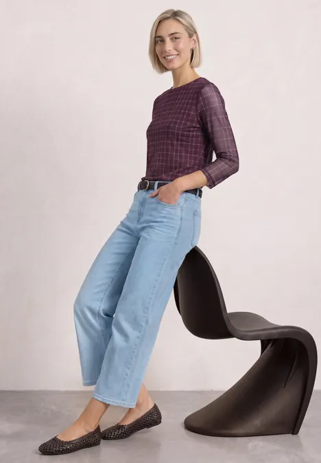 Shirt met 3/4-mouwen van mesh met print rustic rouge Eine lächelnde Frau in karierter Bluse, lockeren Jeans und schwarzen Ballerinas, lehnt an einem modernen Stuhl.