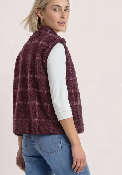 Frau im lila karierten Fleece-Weste über weißem Langarmshirt und blauen Jeans, lächelnd über die Schulter blickend.