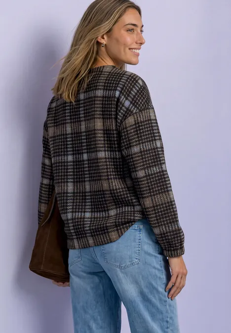 Frau in kariertem Pullover und Jeans schaut lachend über die Schulter, hinter ihr ist eine helle Wand.