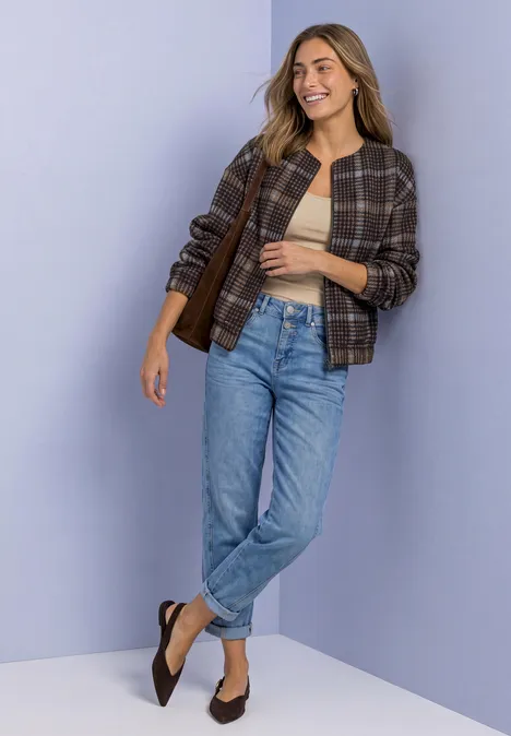 Frau in Jeans, beigem Top und kariertem Blouson, lächelnd vor hellem Hintergrund.