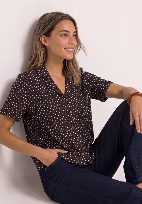 Shirt met verschillende texturen en een knoopsluiting black coffee Een vrouw zit ontspannen, draagt een zwart-wit gestipte blouse met een donkere broek en lacht.