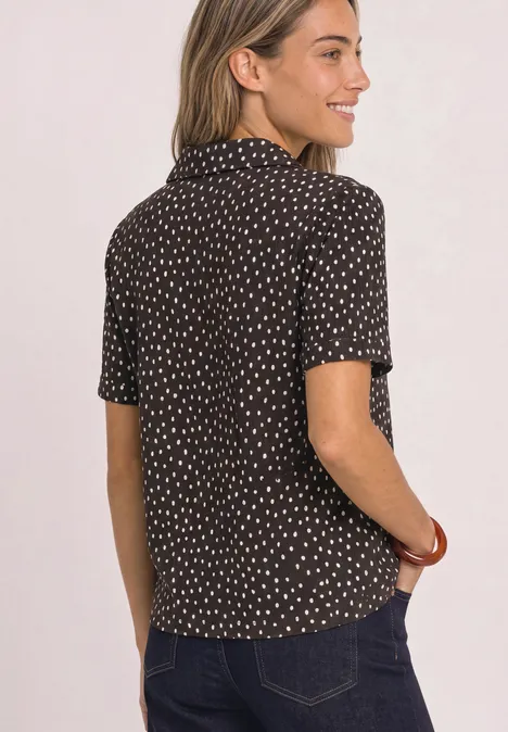 Shirt met verschillende texturen en een knoopsluiting black coffee Vrouw van achteren in een zwarte blouse met witte stippen en spijkerbroek, glimlachend zijwaarts kijkend.