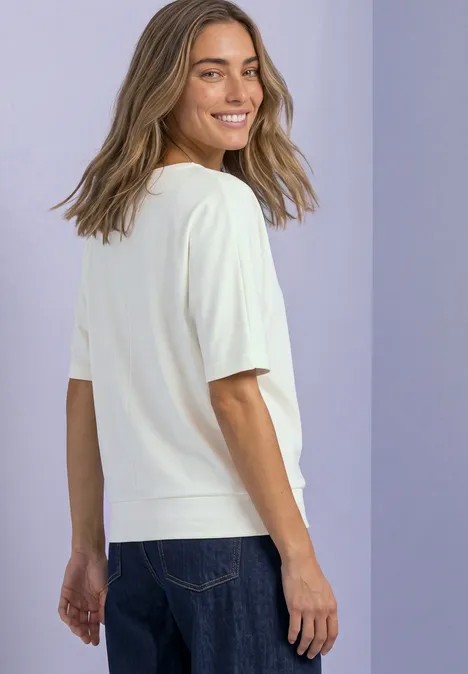 Frau in weißem T-Shirt und Jeans lächelt über die Schulter vor lila Hintergrund.