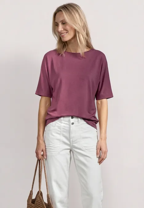 Eine Frau steht vor neutralem Hintergrund, trägt ein violettes T-Shirt, weiße Jeans und hält eine braune Tasche.