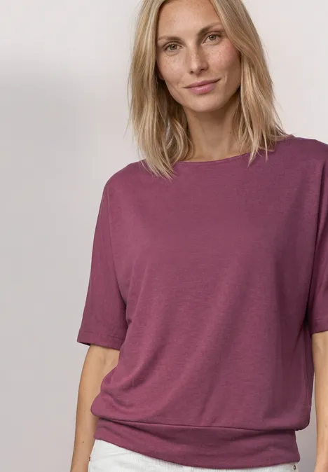Eine Person trägt ein locker sitzendes, pflaumenfarbenes T-Shirt mit kurzen Ärmeln vor hellem Hintergrund.
