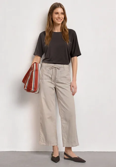 Frau in schwarzem T-Shirt, beigen weiten Hosen, schwarzen Flats und rotem gestreiften Shopper.