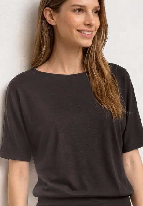 Frau mit schulterlangem braunem Haar trägt ein schlichtes schwarzes T-Shirt vor hellem Hintergrund.