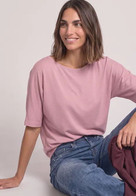 Eine Frau sitzt lächelnd, trägt ein rosafarbenes T-Shirt und Jeans vor neutralem Hintergrund.