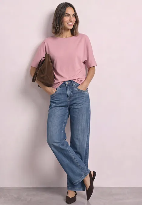 Frau in rosa T-Shirt, blauen Jeans und schwarzen Schuhen steht lächelnd und trägt eine braune Tasche.
