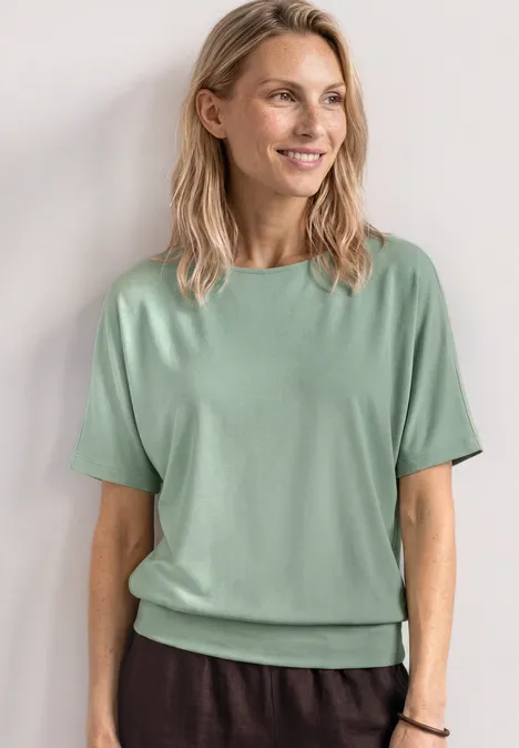 Frau in mintgrünem T-Shirt und dunklen Hosen vor hellem Hintergrund.