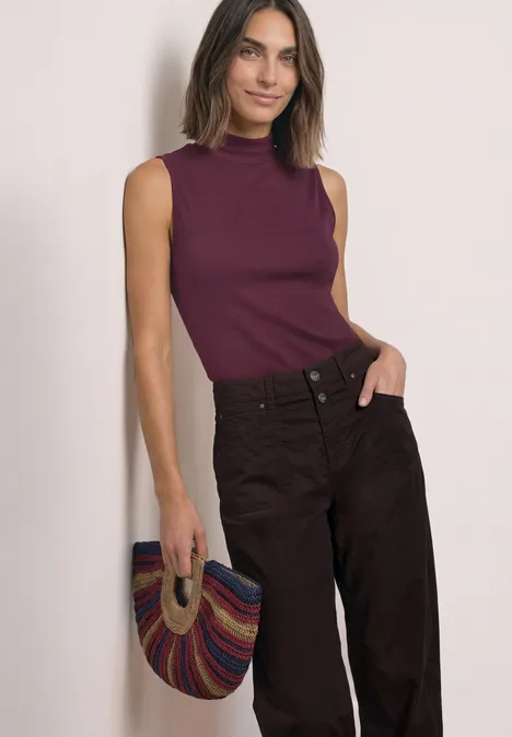 Top im Silk-Look mit Turtle Neck rustic rouge Frau in ärmellosem beerenfarbenem Top und dunkler Hose, hält eine bunte, gestreifte Tasche.