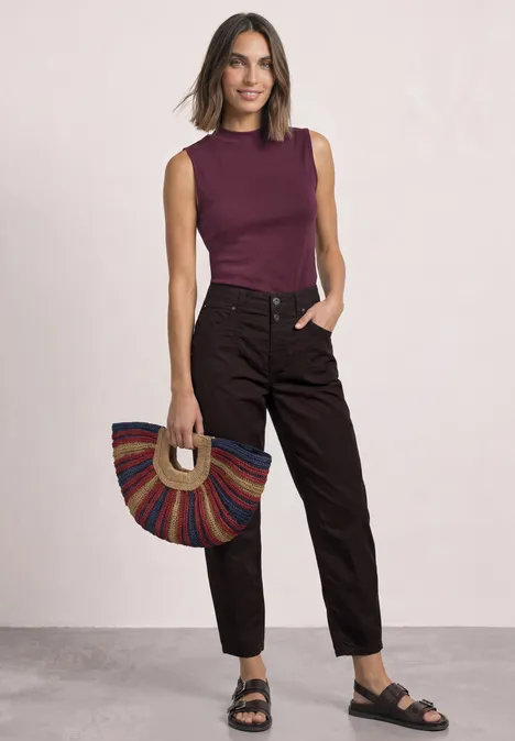 Top im Silk-Look mit Turtle Neck rustic rouge Frau in ärmellosem bordeauxfarbenem Oberteil, dunklen Bundfaltenhosen und schwarzen Sandalen mit bunter, halbrunder Tasche.