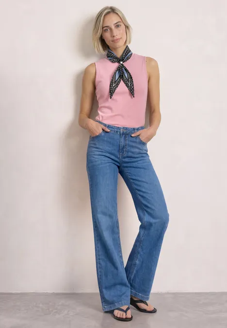 Eine Frau in pinkem Top und weiten Jeans steht vor hellem Hintergrund mit Händen in den Hosentaschen.