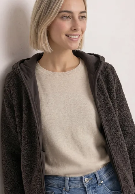 Junge Frau in Jeans, beige Pullover und brauner Teddy-Fleecejacke mit Kapuze vor hellem Hintergrund.