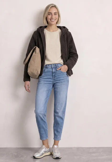 Frau in lässiger Kleidung: braune Jacke, weißes Shirt, Jeans, silberne Sneaker und Tasche vor heller Wand.