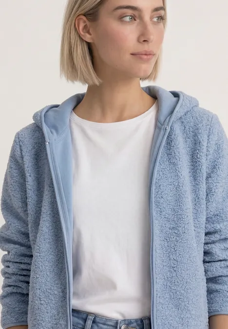 Junge Frau mit kurzem blonden Haar in hellblauem Fleece-Hoodie und weißem T-Shirt vor neutralem Hintergrund.