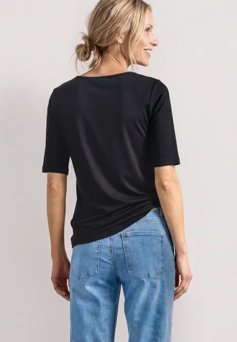 Rückansicht einer Frau in schwarzem T-Shirt und blauer Jeanshose vor hellem Hintergrund.