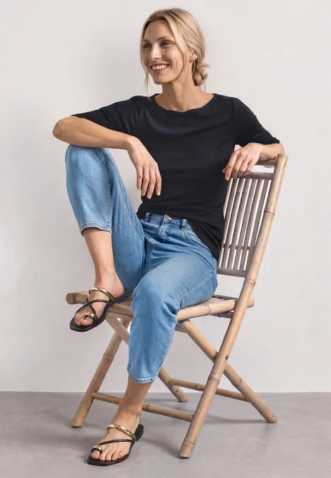 Person sitzt auf Holzklappstuhl, trägt schwarzen Pullover, blaue Jeans und Sandalen.