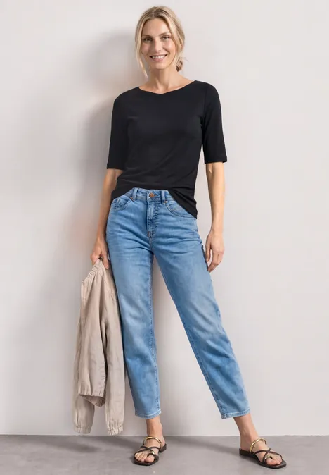 Frau mit schwarzem T-Shirt, hellblauen High-Waist-Jeans, schwarzen Sandalen, hält beige Jacke.