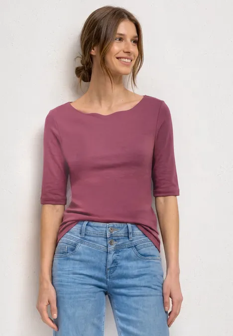 Frau in rosafarbenem Oberteil mit halblangen Ärmeln und blauen Jeans vor weißem Hintergrund.