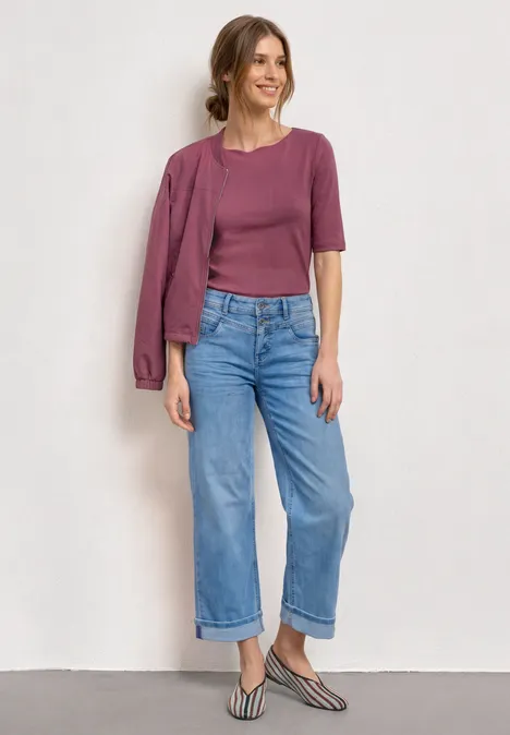 Eine Frau in lila Top und Jacke über der Schulter trägt weite Jeans und gestreifte Slingback-Schuhe.