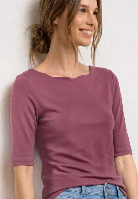 Eine Frau in einem rosafarbenen T-Shirt mit halblangen Ärmeln vor weißem Hintergrund.