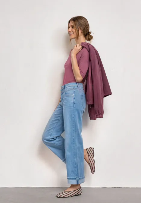 Frau lehnt lächelnd an weißer Wand, trägt pinkes Top, lila Jacke, weite blaue Jeans und gestreifte Slipper.