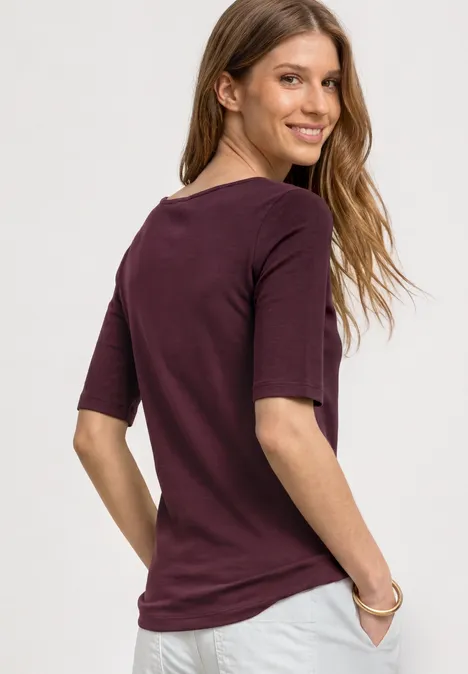 Junge Frau von hinten in bordeauxfarbenem T-Shirt mit halblangen Ärmeln, Hände in den Taschen.