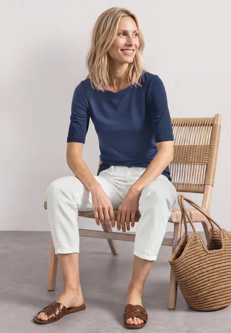 Femme assise sur une chaise, portant un t-shirt bleu, un pantalon blanc, des sandales marron et un sac tressé.