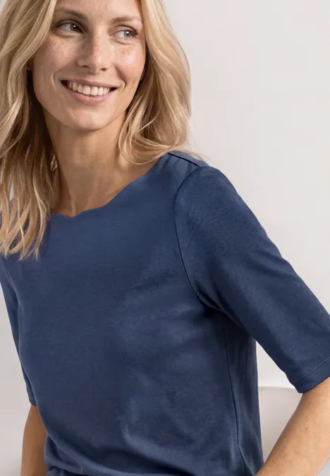 Une femme aux cheveux blonds porte un haut bleu foncé à manches mi-longues sur fond blanc.