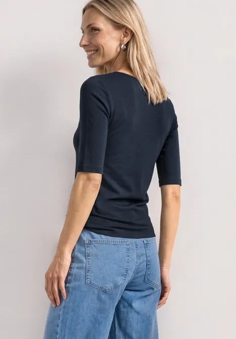 Eine Frau von hinten in dunkelblauem Kurzarmshirt und hellblauen Jeans vor hellem Hintergrund.
