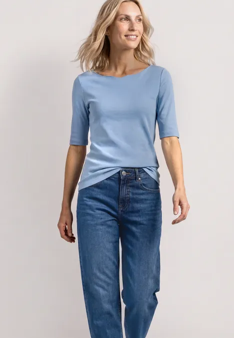 Eine Frau steht in einem hellblauen T-Shirt und blauen Jeans vor einem neutralen Hintergrund.