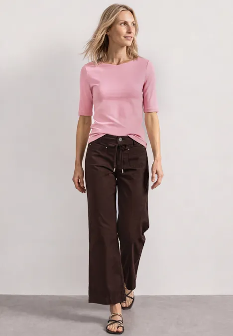 Femme en haut rose et pantalon marron ample se tient devant un fond neutre.