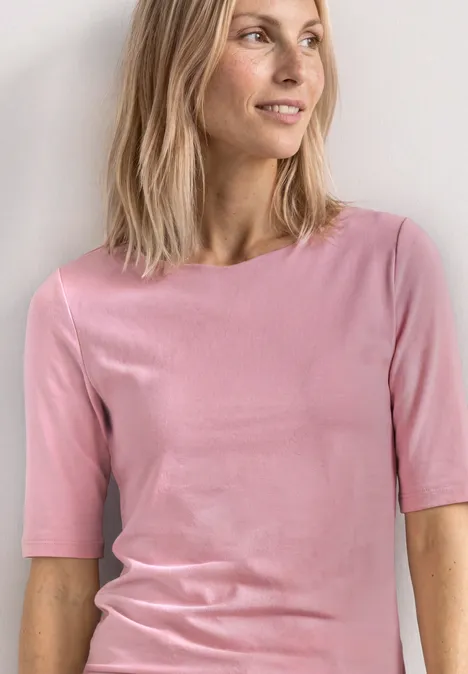 Personne porte un t-shirt rose cintré et cheveux mi-longs devant un fond clair.