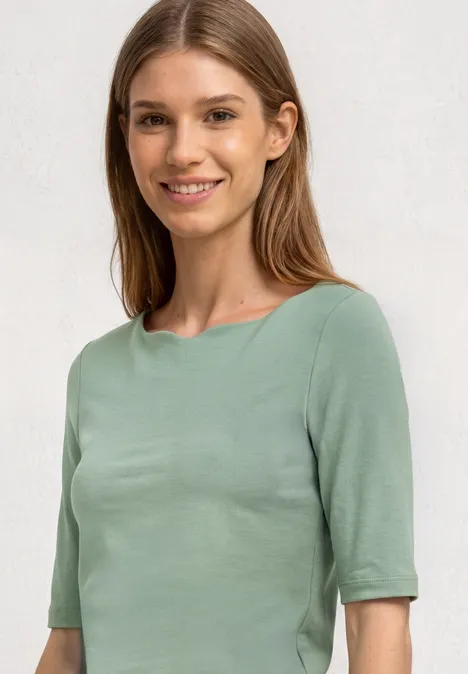 Eine Frau trägt ein schlichtes, hellgrünes Kurzarmshirt vor hellem Hintergrund.