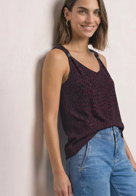 Top mit V-Neck und Dekodetail rustic rouge Eine lächelnde Frau lehnt an einer Wand, trägt ein ärmelloses, dunkelrotes gemustertes Top und blaue Jeans