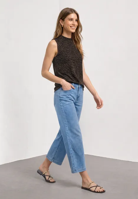 Eine Frau in einem ärmellosen, schwarz gemusterten Top, blauen Jeans und flachen, schwarzen Sandalen.