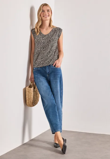 Shirt mit Boat Neck und Elastikbund cotton beige Frau lehnt an Wand, trägt Leo-Top, blaue Jeans, schwarze Ballerinas und hält eine Strohtasche.