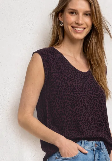 Frau im ärmellosen, dunkelroten Leoparden-Top und Jeans vor weißer Wand.